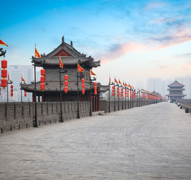 Historical building standing along ancient city wall, adorned with red lanterns and flags. Surrounded by clear blue sky with soft clouds, the path stretches into the distance, flanked by more structures.