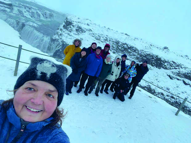 A person smiles for a selfie in snowy, winter clothing, with a group of people behind them standing on a snow-covered landscape, featuring a distant waterfall and mountainous terrain.