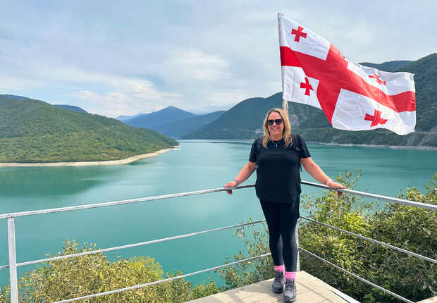 A woman stands smiling on a platform by a blue lake surrounded by mountains, holding a railing. A large Georgian flag flutters beside her under a cloudy sky.