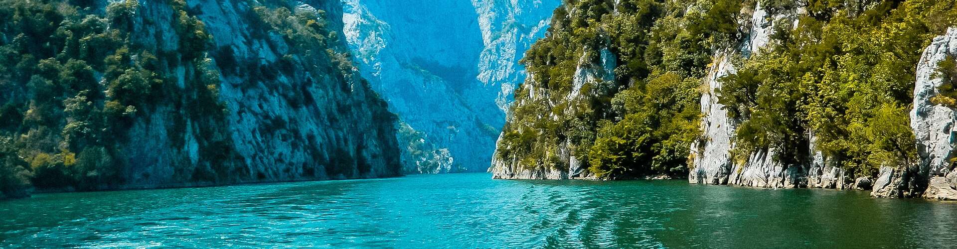 Turquoise river flows between steep, forested cliffs, under a bright blue sky, creating a serene, natural canyon landscape.