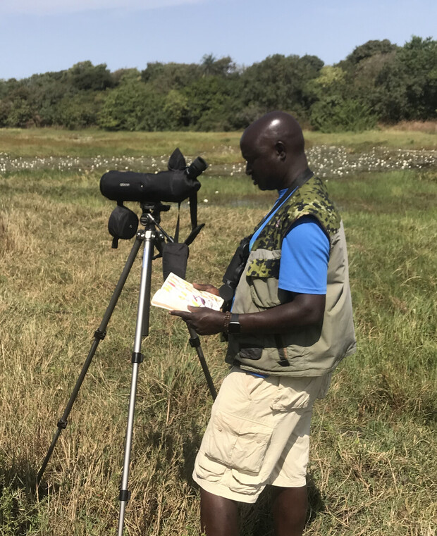 A person examines a field guide while standing next to a spotting scope on a tripod in a grassy area, with trees in the background.