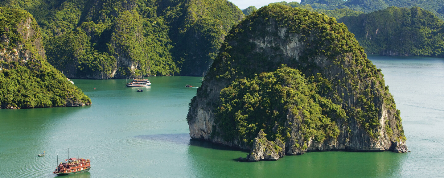 Lush, green limestone islands rise dramatically from calm turquoise waters, while boats smoothly glide between them, set against a backdrop of verdant hills and a clear sky.