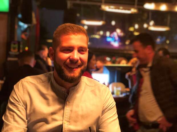 A person with a beard smiling, wearing a light-colored shirt, seated in a lively, dimly lit bar or restaurant. Blurred people and hanging lights create a warm, social atmosphere.