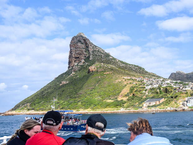 A boat sails near a steep, green-covered mountain, with several people in the foreground observing. The background features scattered buildings along the shoreline under a partly cloudy sky.