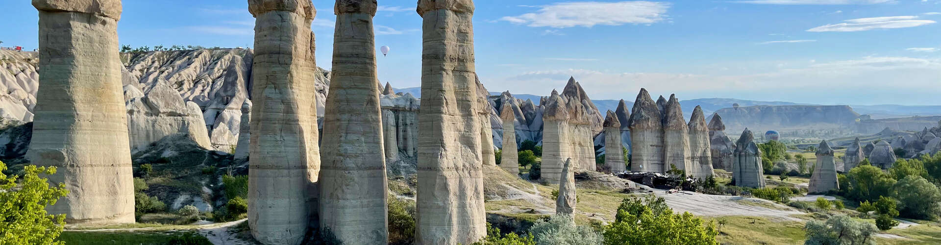 Tall, uniquely shaped rock formations stand amidst a landscape of rolling hills and sparse greenery. Hot air balloons float in the distant sky, enhancing the scenic panorama.