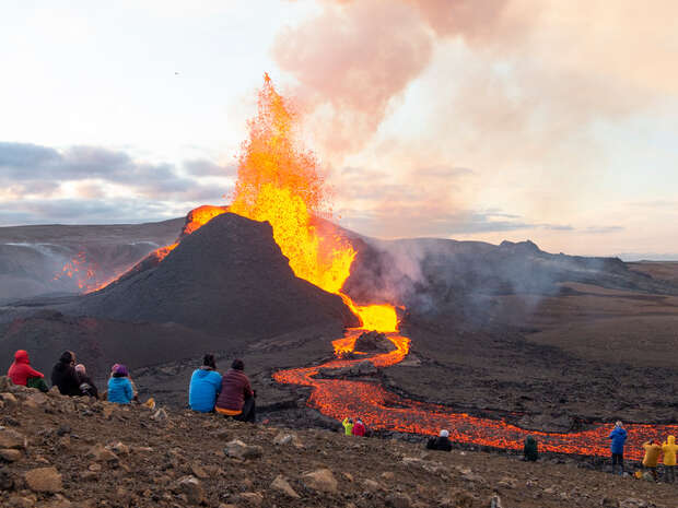 A volcanic eruption spews glowing lava and ash into the sky, flowing down a rocky slope. Nearby, people sit on a ridge observing the scene with barren, mountainous terrain surrounding them.