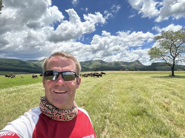 A person wearing sunglasses stands in a grassy field, smiling with a herd of cows in the background. Mountains and a tree line the horizon under a partly cloudy sky.