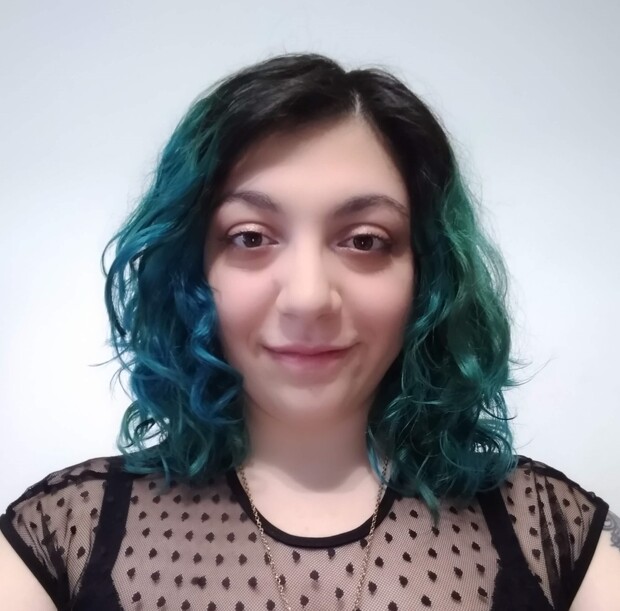 Person with shoulder-length, curly teal hair is smiling slightly, wearing a black, sheer, polka-dotted top. The background is a plain, light-colored wall.