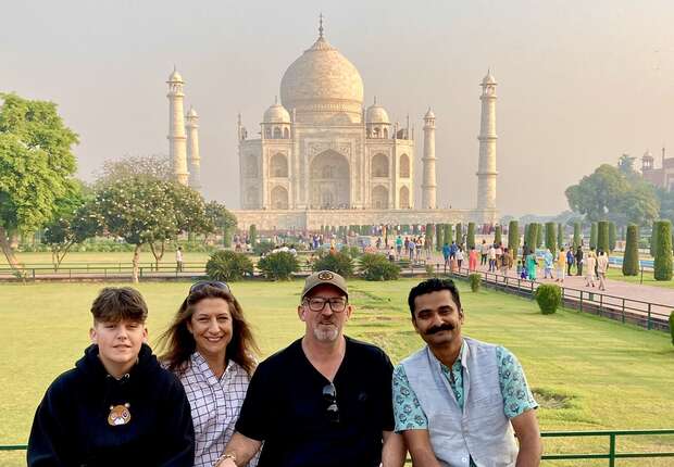 Four people sit together in front of the Taj Mahal, an iconic white marble mausoleum, surrounded by gardens and visitors exploring the area on a sunny day.