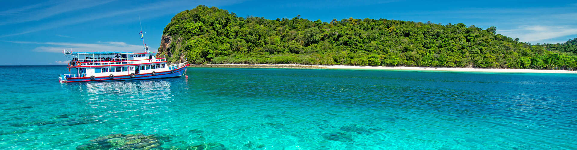 A colorful boat floats on clear, turquoise water near a green, forested island with a sandy beach under a bright, blue sky.