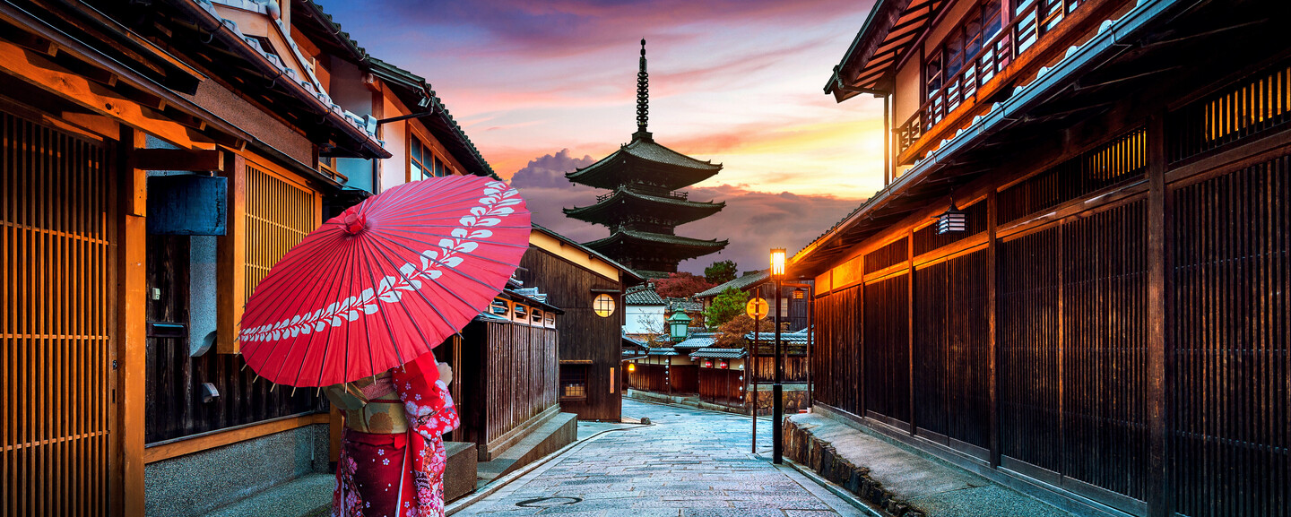 A person in traditional attire holds a red parasol, walking through a narrow, historical Japanese street at sunset, with a pagoda visible in the distance.