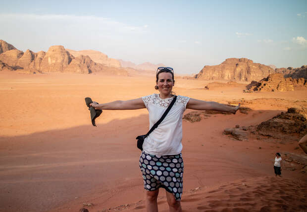 A person stands with arms outstretched, smiling, holding sunglasses on a vast, rocky desert landscape with distant hills under a clear sky.