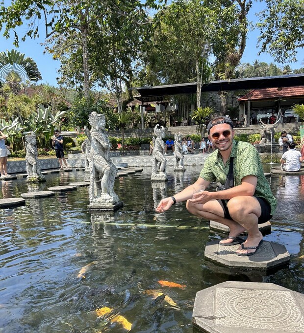 A person squats on a stepping stone in a pond, feeding koi fish. Statues and tourists surround the water, with tropical plants and a building in the background.