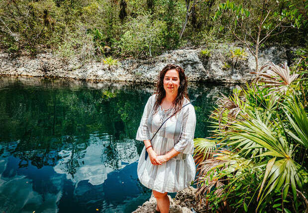 A person stands smiling by a clear blue cenote, surrounded by lush green foliage and rocky terrain, under a bright, sunny sky.