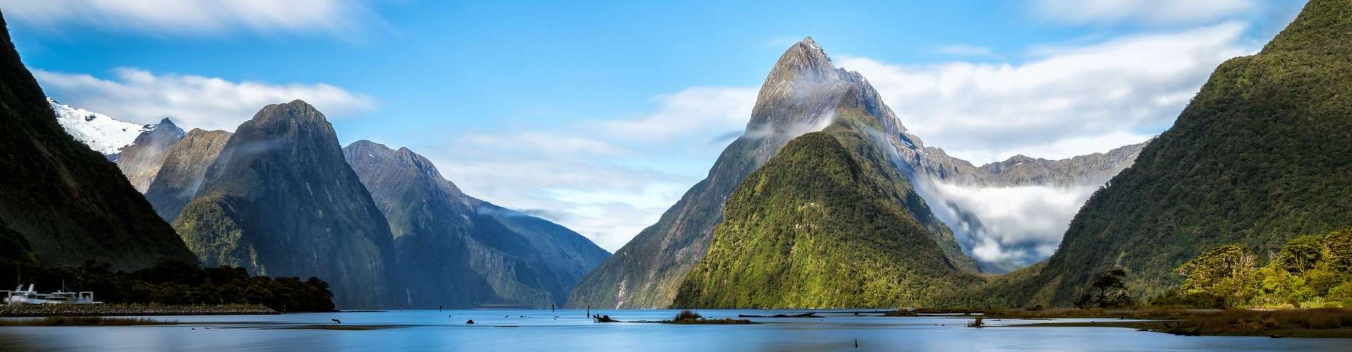 Snow-capped mountains rise majestically, draped in clouds, beside a serene blue lake surrounded by lush green forests. The sky is a bright blue with scattered white clouds.