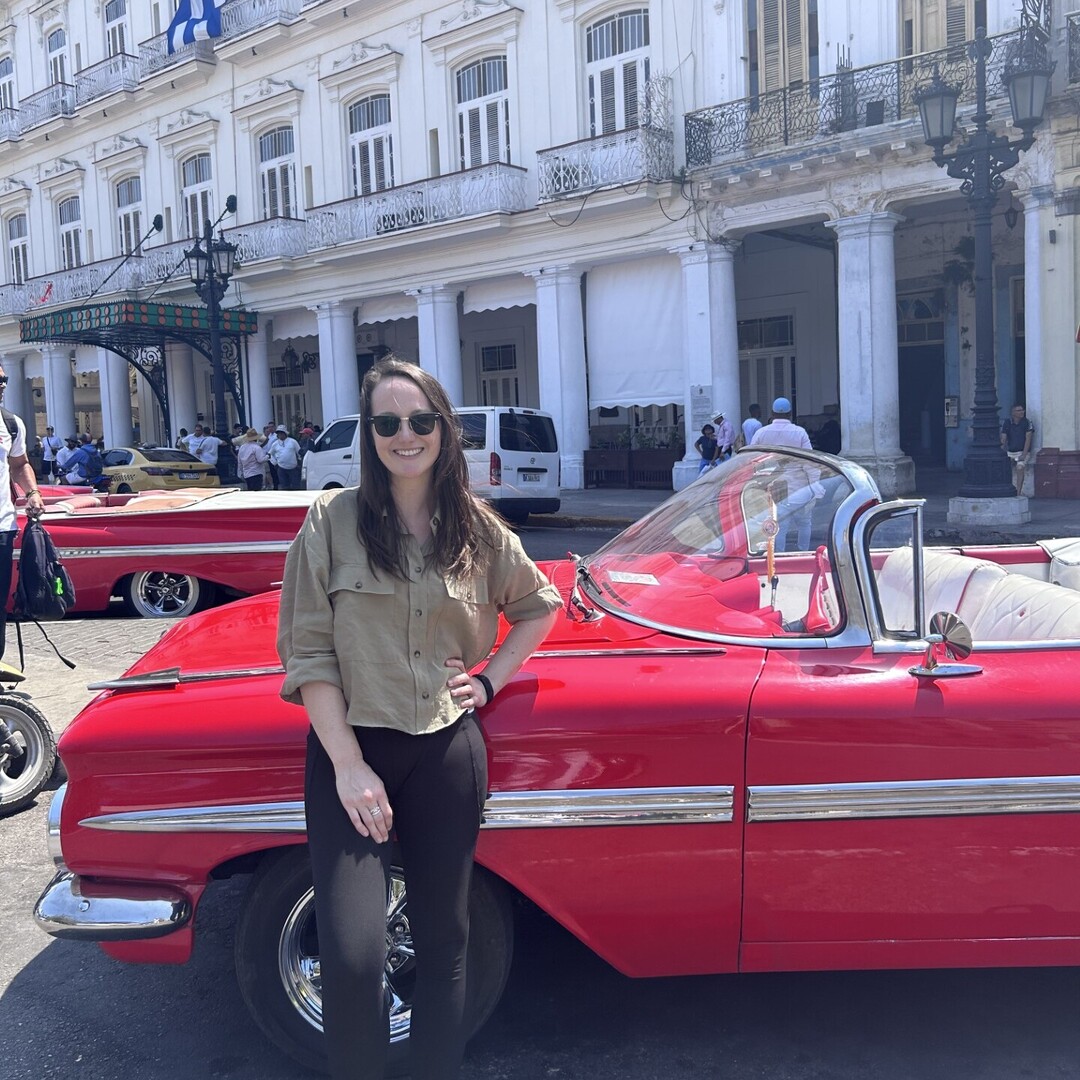A person stands smiling beside a bright red vintage car, parked in front of a white, historic building with arched windows and columns. Other classic cars and people are visible nearby.