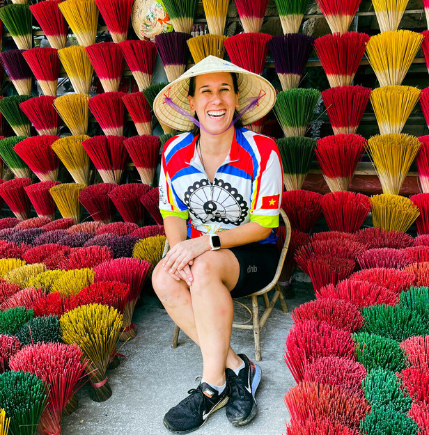 Person sitting on a stool, smiling, wearing a conical hat and colorful shirt. Surrounded by vibrant bundles of incense sticks in red, yellow, and green, creating a bright, artistic backdrop.