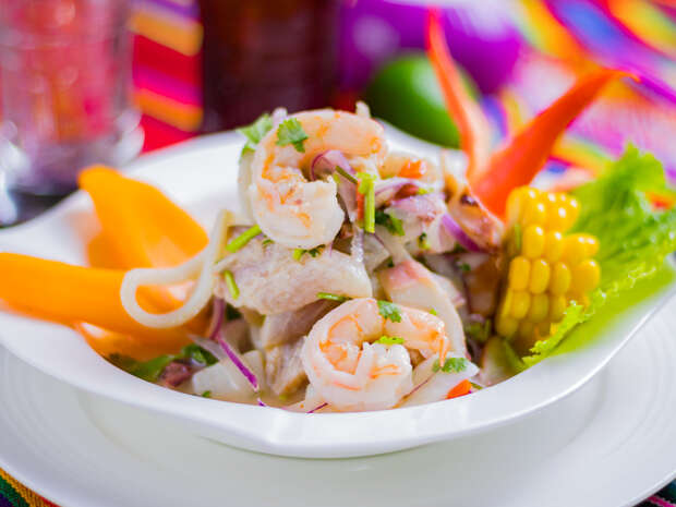 Shrimp ceviche sits in a white bowl, garnished with corn, orange bell peppers, and lettuce. It rests on a colorful tablecloth that adds vibrant context to the dish.