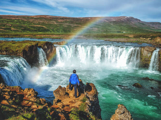 A person in a blue jacket stands on rocky terrain, overlooking a wide waterfall beneath a rainbow, surrounded by lush greenery and distant hills under a partly cloudy sky.
