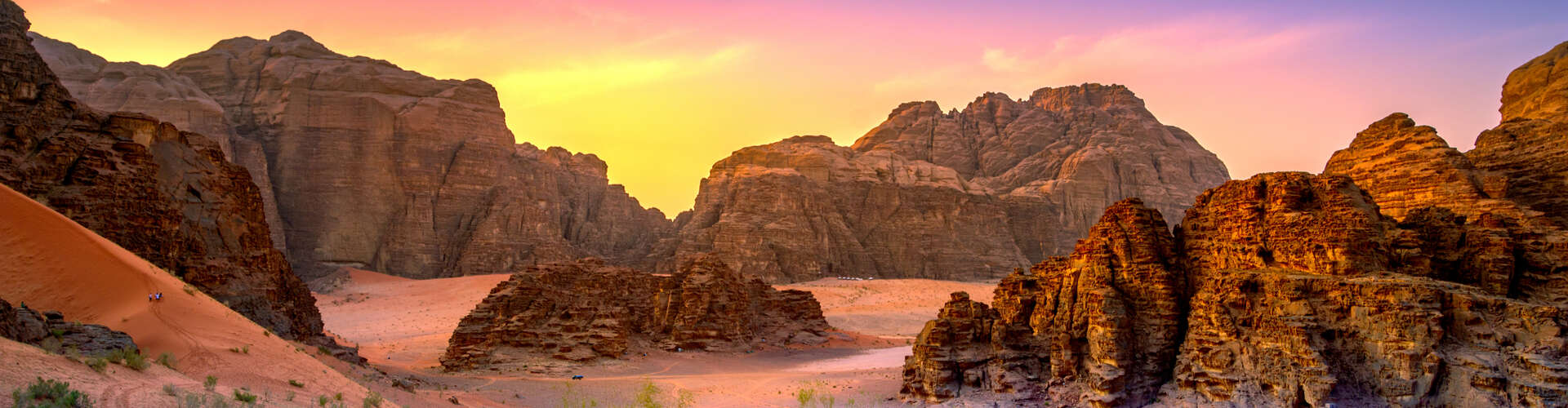Rocky cliffs frame two small figures walking across a reddish sand dune at sunset, with a vibrant sky illuminating the rugged desert landscape.