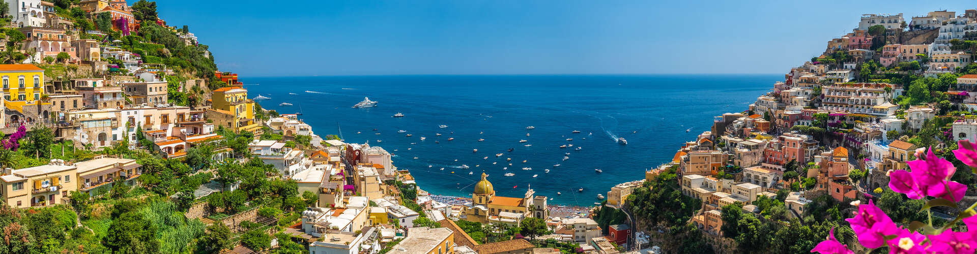 Colorful hillside buildings cascade down to the sea, with numerous small boats dotting the blue water. Lush greenery and vibrant flowers frame the vibrant townscape under a clear sky.
