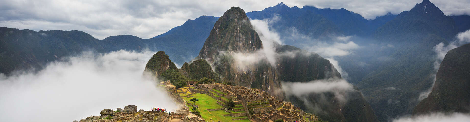 Ruins sit atop a mountain ridge, partially obscured by clouds, with steep, surrounding mountains under a cloudy sky, conveying an ancient, mystical atmosphere.