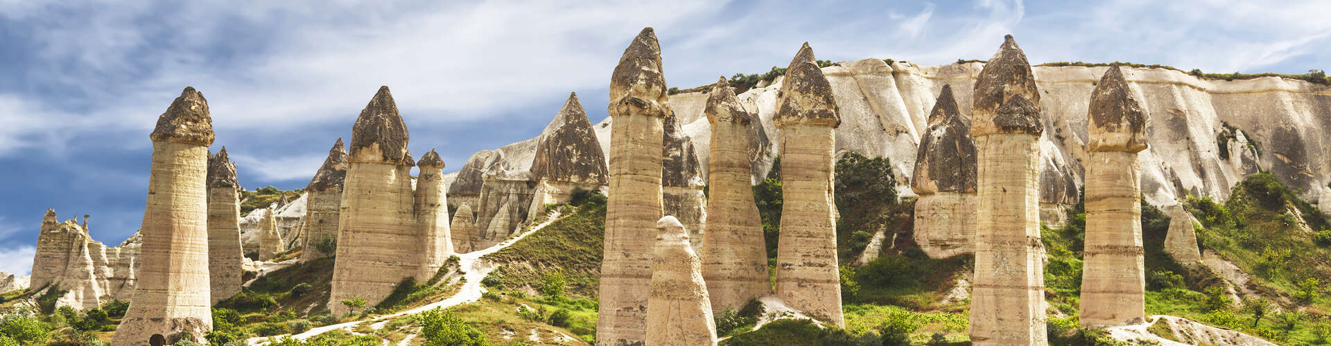Tall, rock formations resembling spires stand prominently in front of rugged cliffs, surrounded by sparse greenery under a partly cloudy sky.
