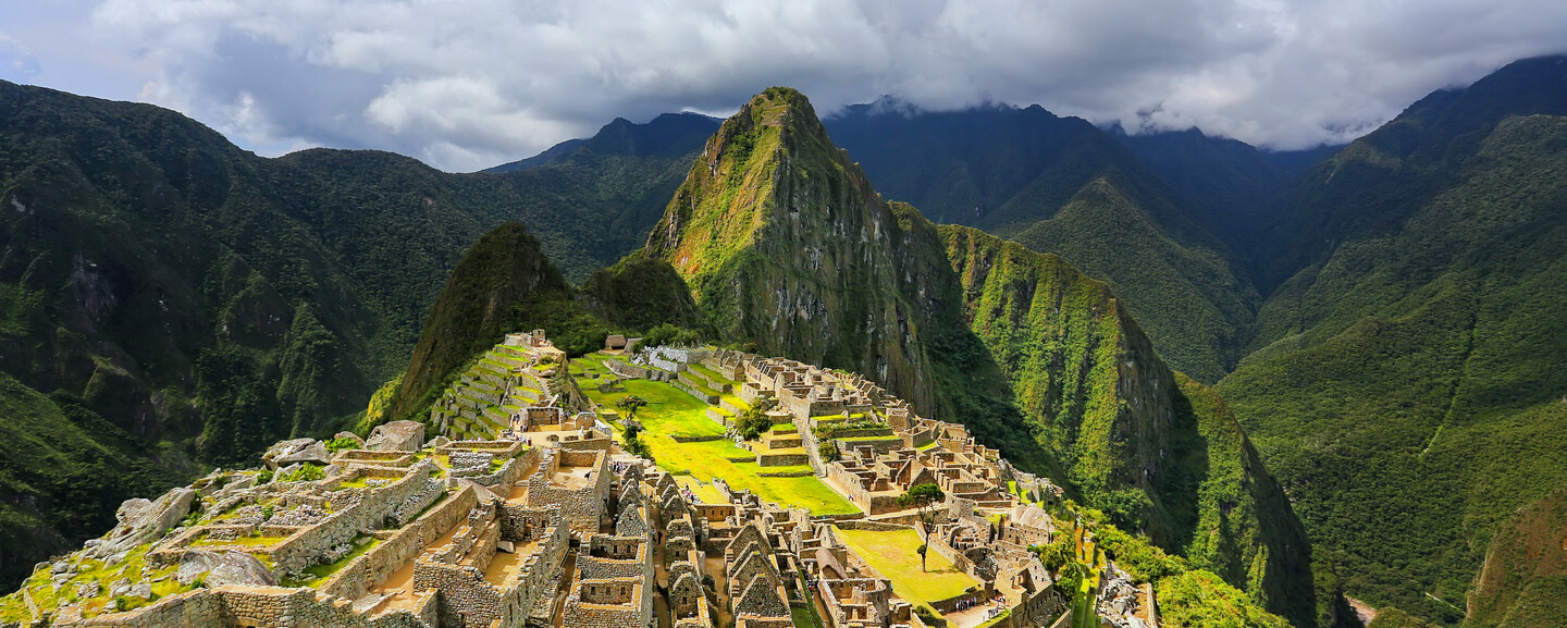 Ancient stone ruins rest on a mountainous slope, surrounded by lush green peaks under a cloudy sky, showcasing a dramatic Andean landscape.