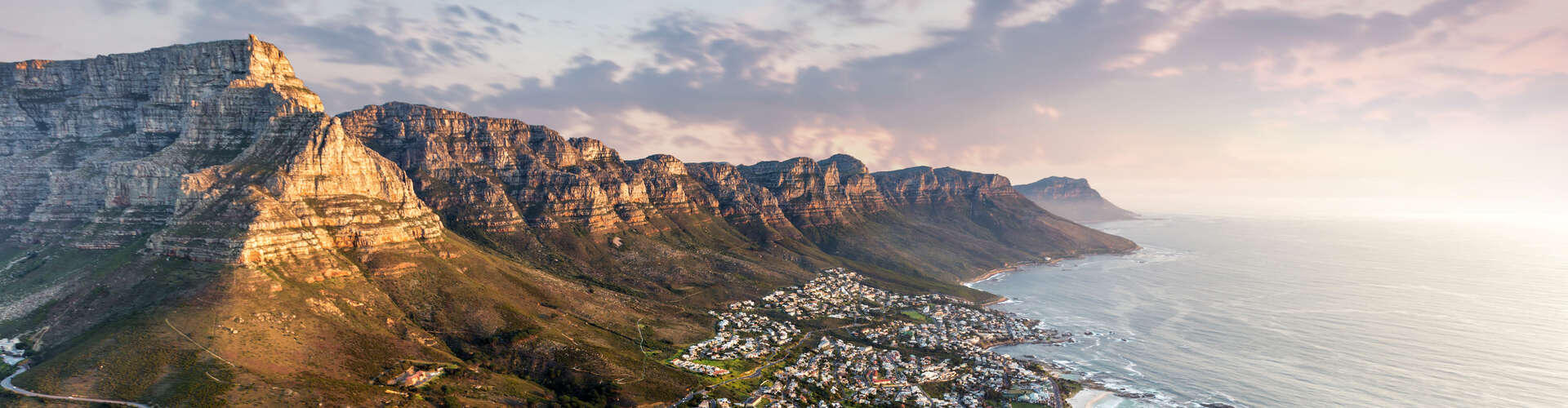 Majestic mountain range bathed in sunlight, towering over a coastal town nestled beside the ocean, with waves gently lapping against the shore under a vibrant, cloudy sky.