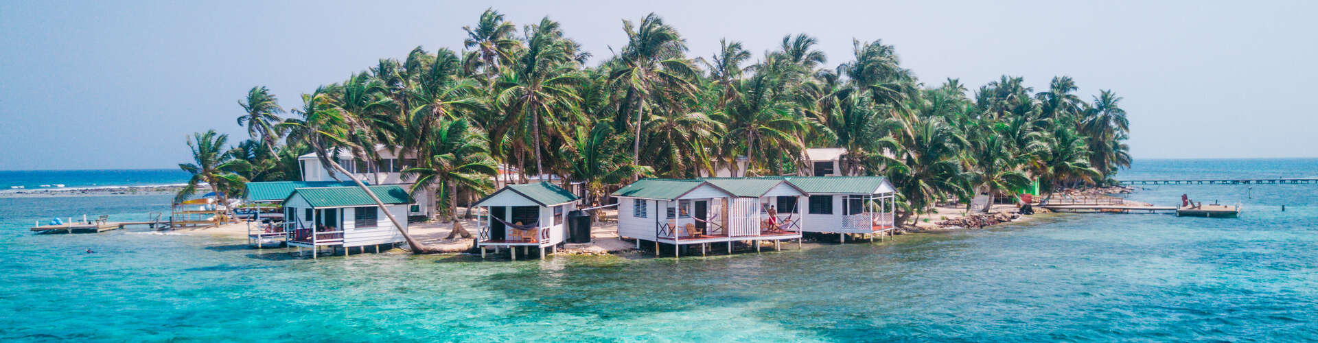Small cottages stand on stilts over clear turquoise water, surrounded by palm trees, creating a tropical island setting with a distant dock and blue sky backdrop.