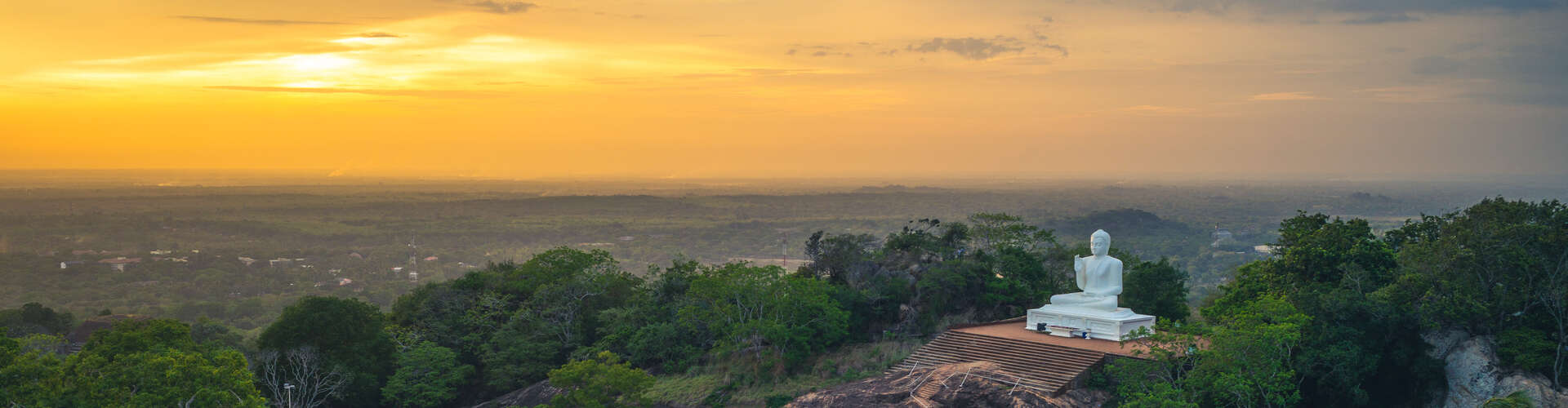 A large white Buddha statue sits serenely on a platform overlooking a vast, green landscape at sunset, with the sky painted in warm orange hues.