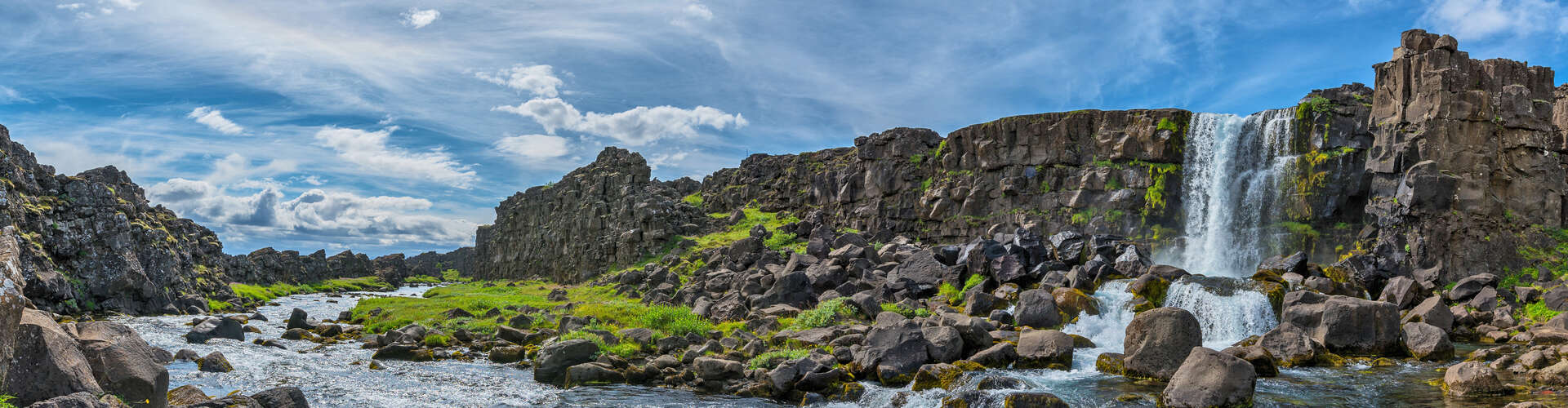 Waterfall cascades down rocky cliff, surrounded by scattered boulders and green foliage; a stream flows through the scene, set against a backdrop of blue sky with wispy clouds.
