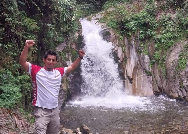 A man stands with arms raised in a triumphant pose near a small waterfall, surrounded by lush green vegetation and rocky terrain. He's wearing a white and red shirt labeled "EXPLORER."