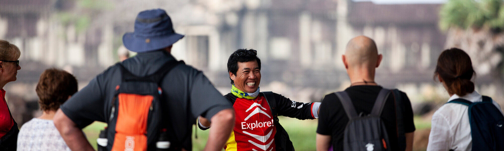 A person wearing an "Explore!" shirt gestures happily while surrounded by a group of people outdoors, with blurred historical architecture in the background.