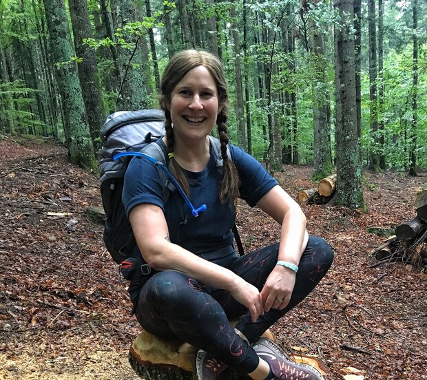 A person with braided hair, wearing hiking gear and a backpack, smiles while crouching on a tree stump. The setting is a lush, green forest with thick tree coverage.