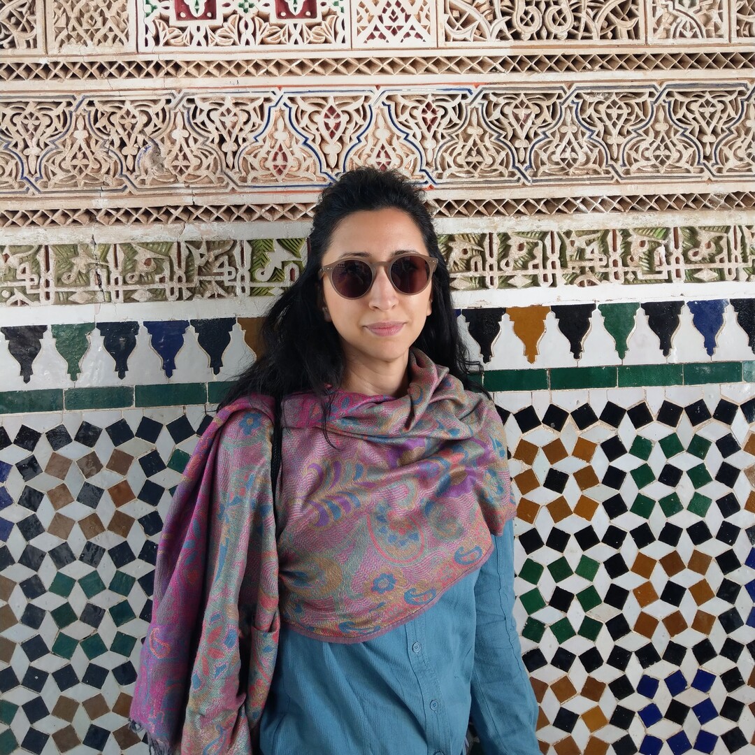 A person wearing sunglasses and a patterned shawl stands in front of a detailed, ornate wall featuring geometric and floral tile designs in various colors.