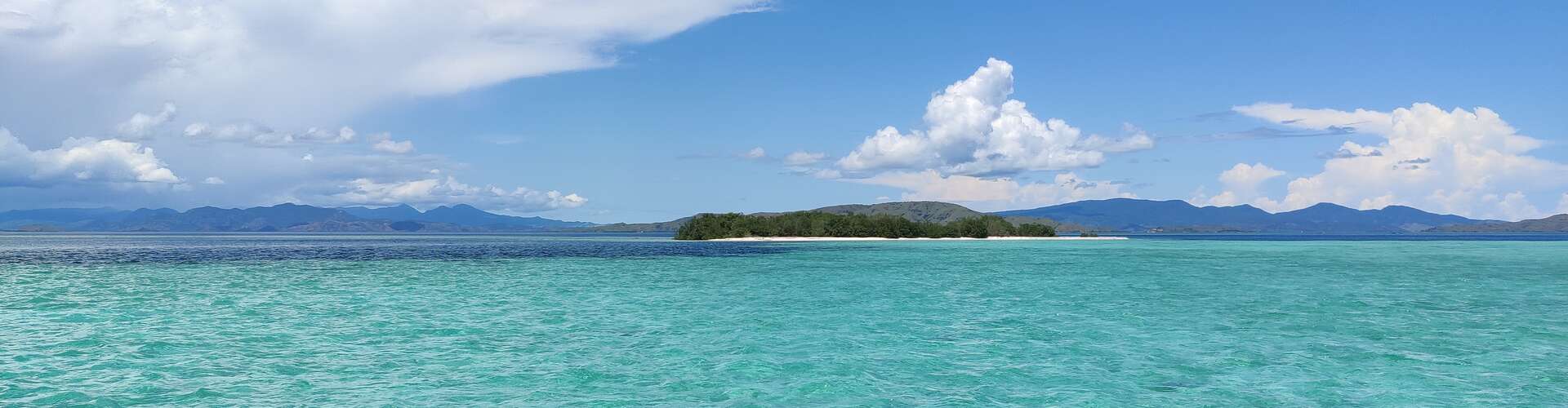An island sits quietly with lush greenery surrounded by turquoise water under a bright blue sky with scattered white clouds. Distant mountains are visible on the horizon.