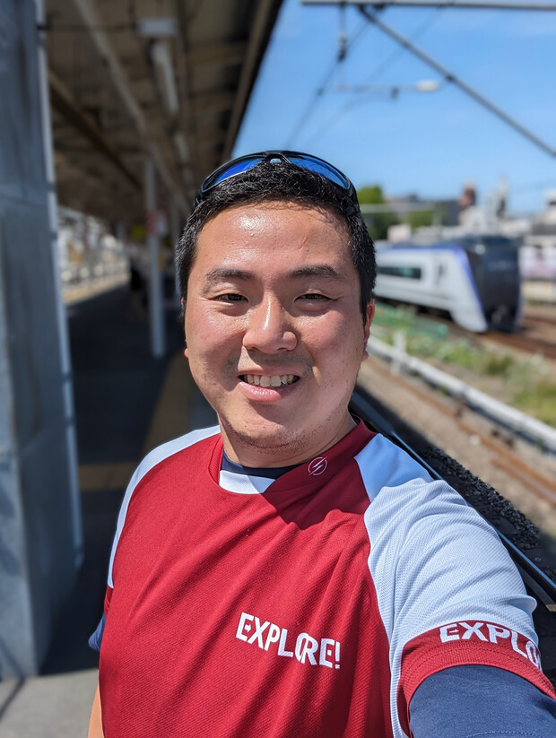 A person wearing a red shirt with "EXPLORE!" written on it smiles at the camera on a sunny train station platform, while a train passes on nearby tracks.