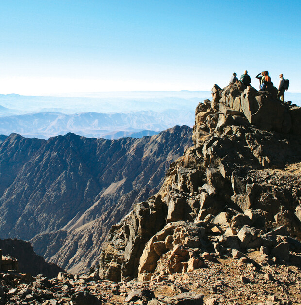 Hikers stand atop a rocky mountain peak, overlooking vast, rugged ridges under a clear blue sky.