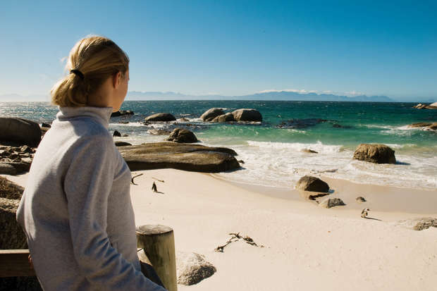 A person wearing a gray sweater gazes at a tranquil beach scene with rolling waves, rocks, and distant mountains under a clear blue sky.