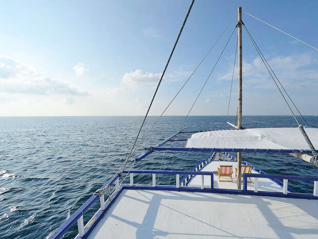 A boat sails on calm ocean water. Wooden deck chairs are positioned on the ship’s sunlit, open deck. A clear blue sky with scattered clouds surrounds the scene.