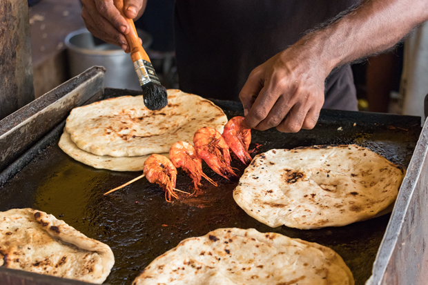 A hand holds skewered shrimp over a hot griddle, applying sauce with a brush. Flatbreads cook nearby, in a busy kitchen setting.