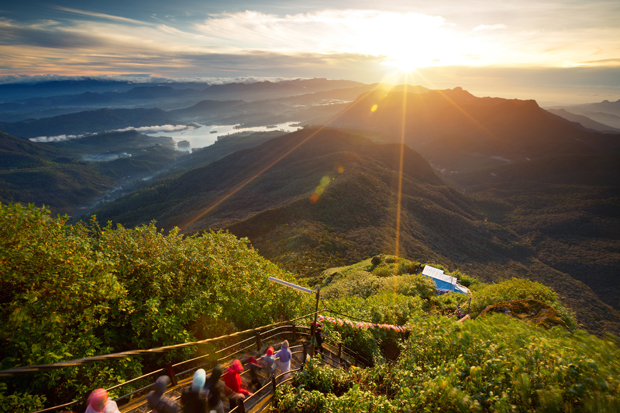 Sun rising over distant mountains, casting golden light over a lush landscape. People are ascending a staircase surrounded by greenery, with a vast valley and winding river below.