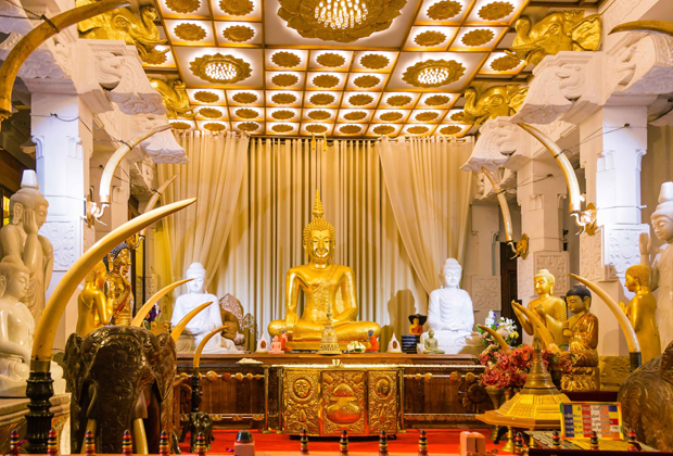 A golden Buddha statue sits serenely on an ornate altar surrounded by white and gold figures, elephant tusks, and decorative elements within a richly adorned temple interior with draped curtains and a patterned ceiling.