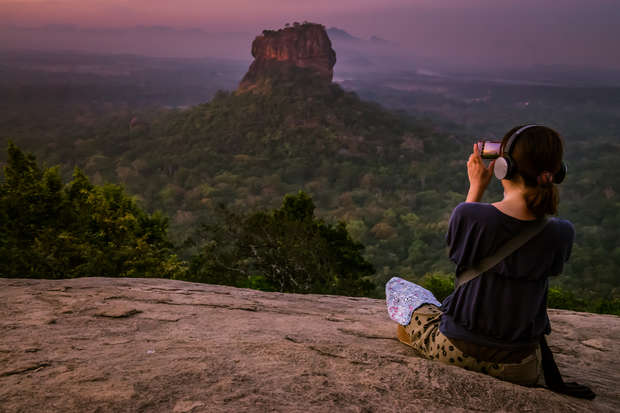 A person sits on a rocky surface, photographing a distant rock formation amidst a lush, expansive forest under a purple-orange sky at sunset. They wear headphones and a bag.