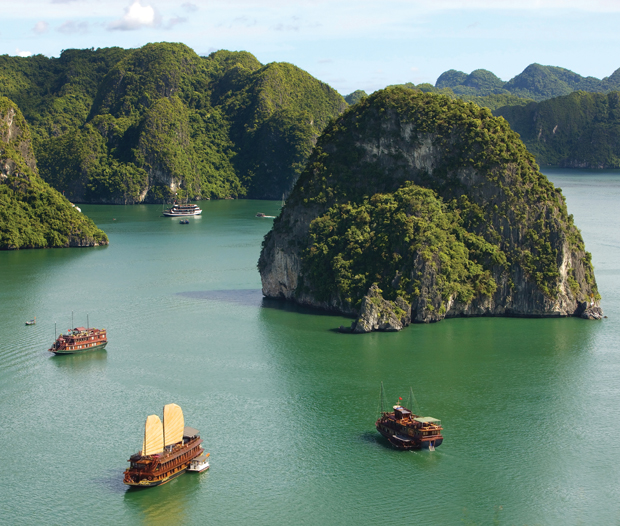 Traditional wooden boats sail through emerald waters surrounded by lush, towering limestone islands and cliffs, under a bright blue sky with scattered clouds.