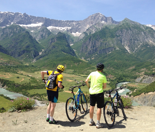 Two cyclists stand beside their bikes, overlooking a scenic valley with lush greenery and a flowing river, set against towering, snow-capped mountains under a clear blue sky.