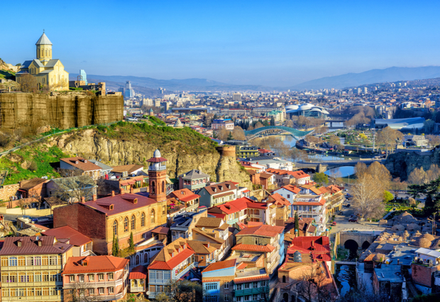 A historical church sits atop a rocky hill, overlooking a vibrant cityscape with colorful buildings and winding rivers, set against distant mountains under a clear blue sky.