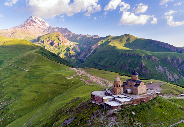 A historic stone church sits atop a grassy hill, overlooking a vast landscape of rolling green mountains, with a towering, snow-capped peak in the background under a cloudy blue sky.