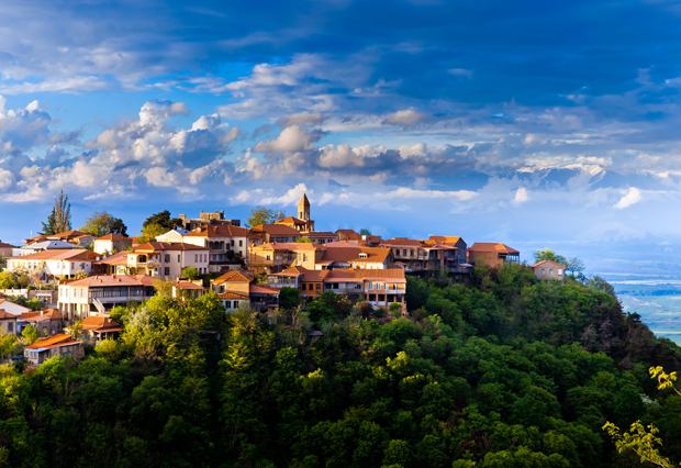 A cluster of rustic buildings with red roofs sits atop a lush, green hill under a dramatic sky filled with billowing clouds, overlooking a distant, expansive landscape.